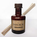 Bangkok Raumduft Diffuser im braunen Glas mit Kraftpapier-Etikett und daneben drapierten Duftstäbchen.